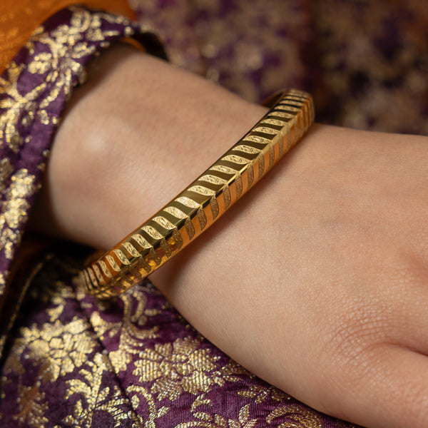 The image shows a gold bangle with a repeating chevron or zigzag pattern. The bangle has a smooth surface and is adorned with the geometric design that runs around its entire circumference. The bangle is worn on a person's wrist, and they are wearing a patterned purple and gold garment.