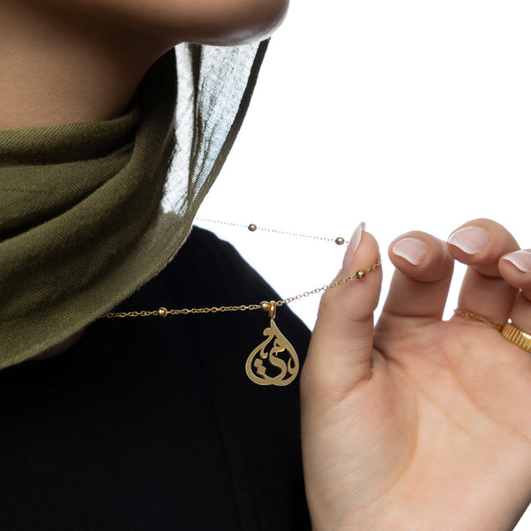 Woman holding the gold Sister Calligraphy Necklace, with visible pendant detailing and beaded chain, styled with a gold ring and olive green hijab.